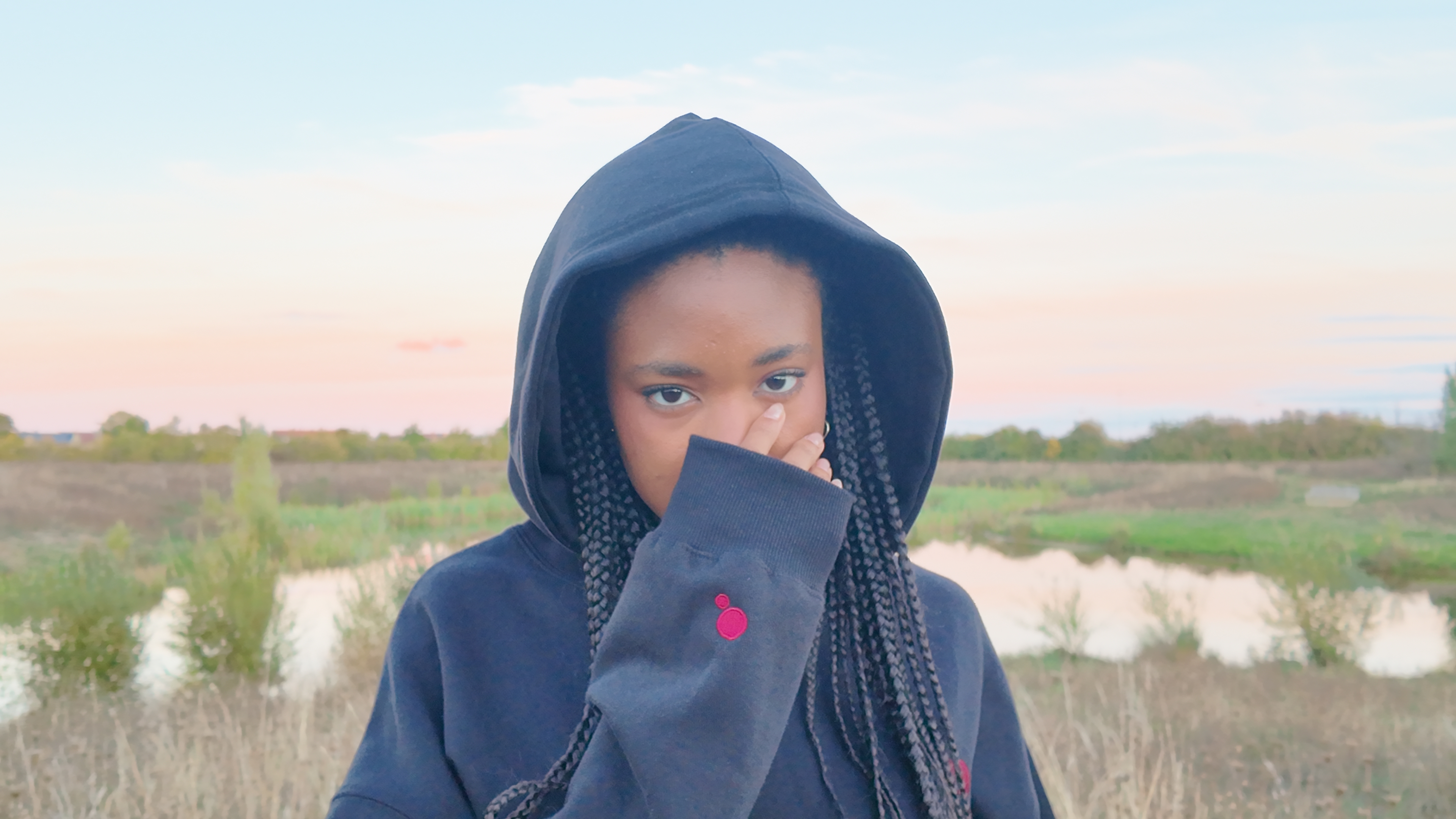 Model in the black Masiix hoodie with the hood up, looking straight at the camera with her hand raised to her face, showing the red dot embroidery on the sleeve