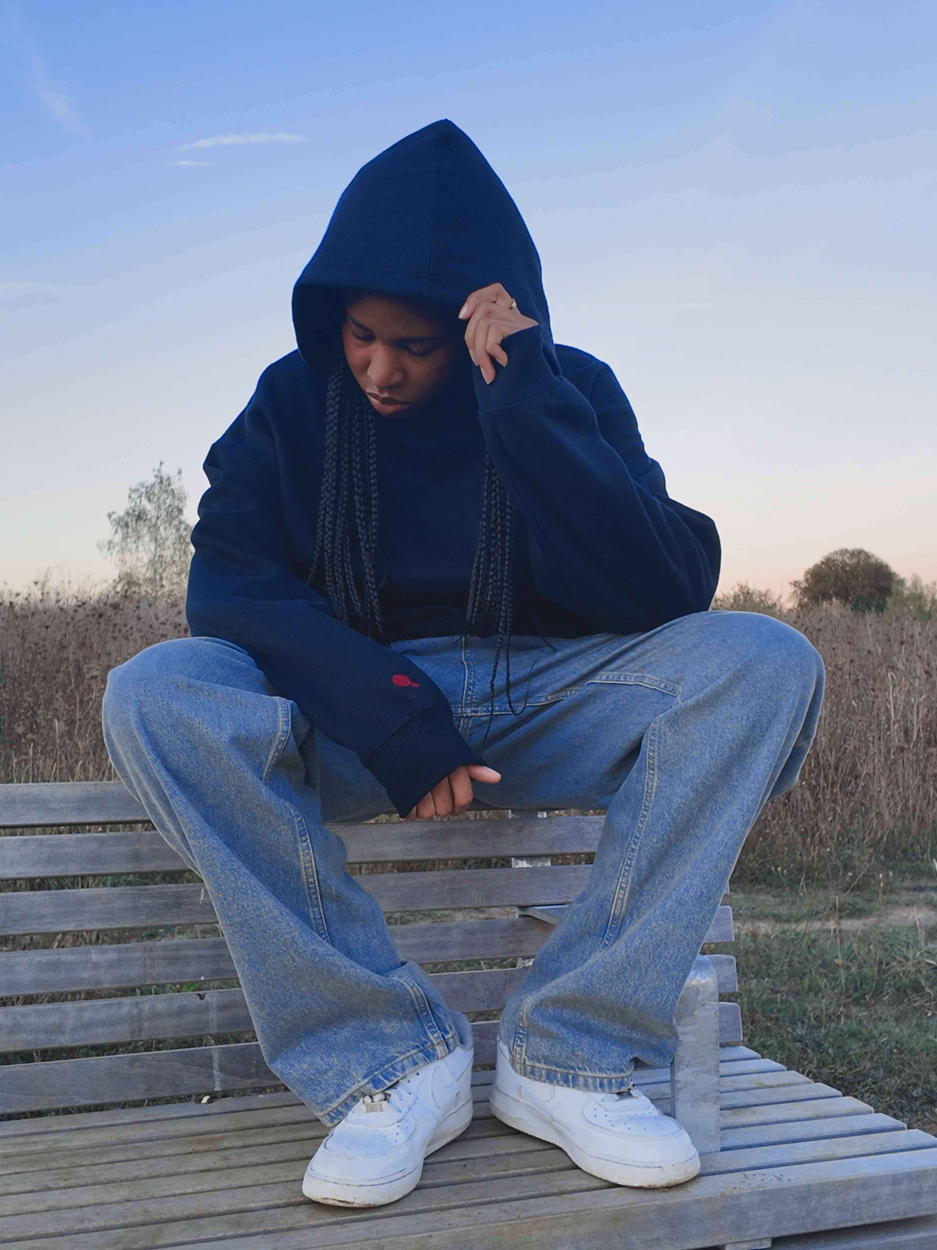 Model seated on a wooden bench, hood up and head bowed in the dark Red Dot Hoodie, the red dot visible on the cuff