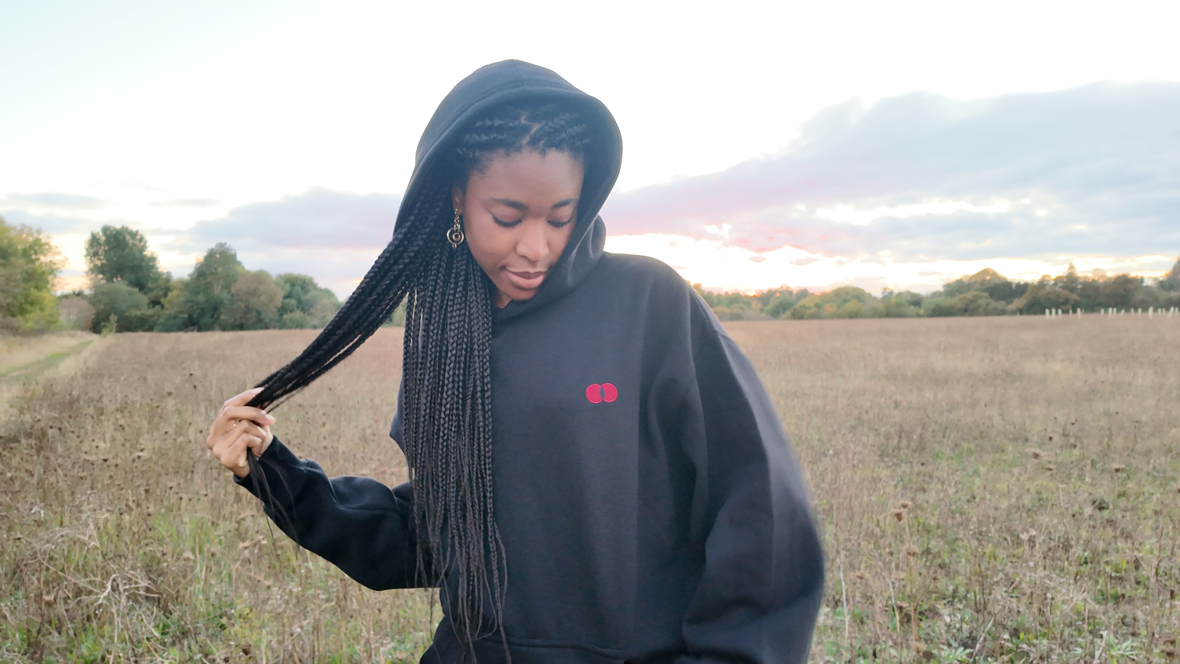 Model in a field at sunset, hood up, looking down and holding her braids, the red double-dot on the chest