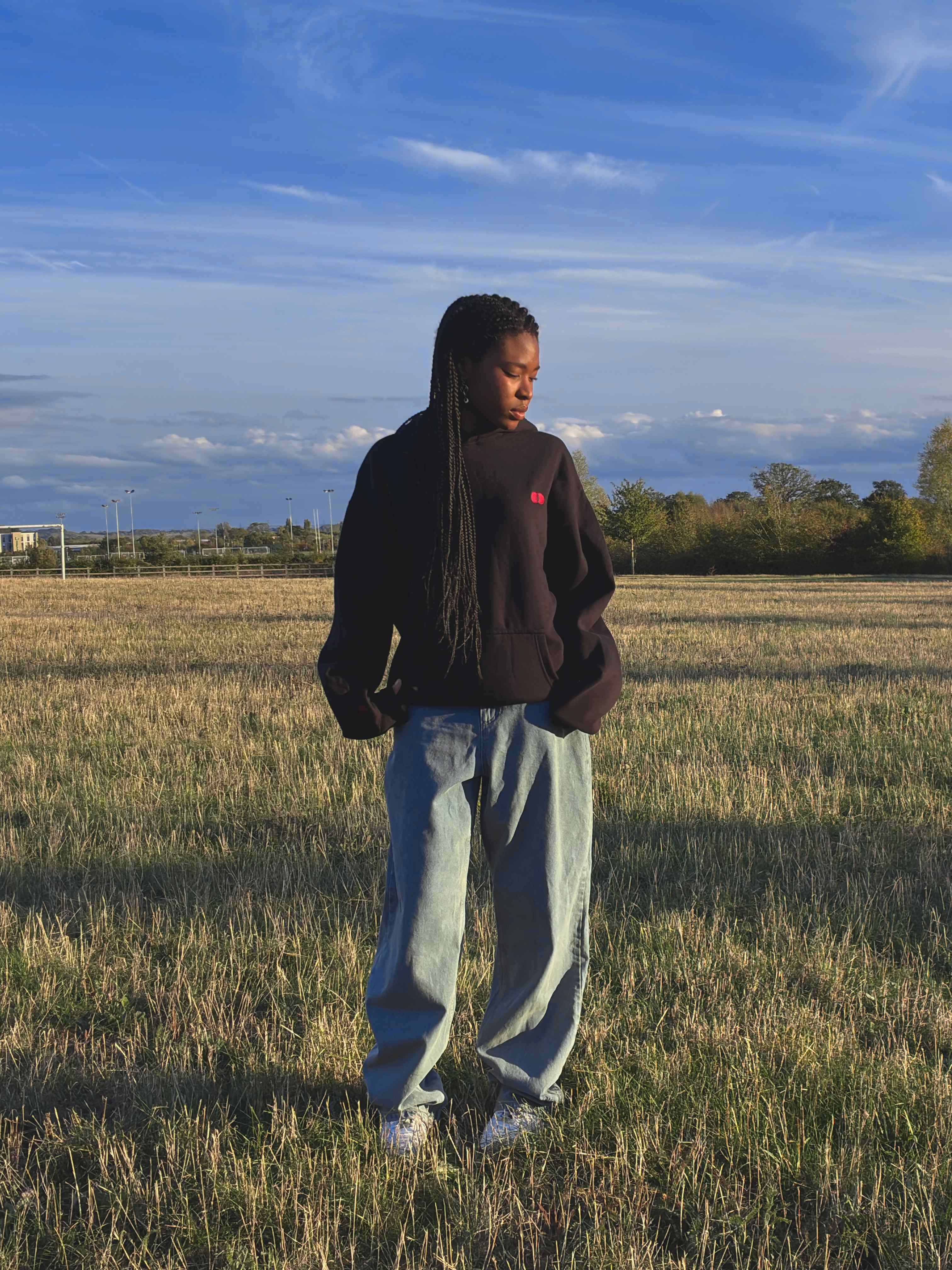 Model standing in a golden field under a blue sky, wearing the black Red Dot Hoodie with light-wash wide-leg jeans and white trainers