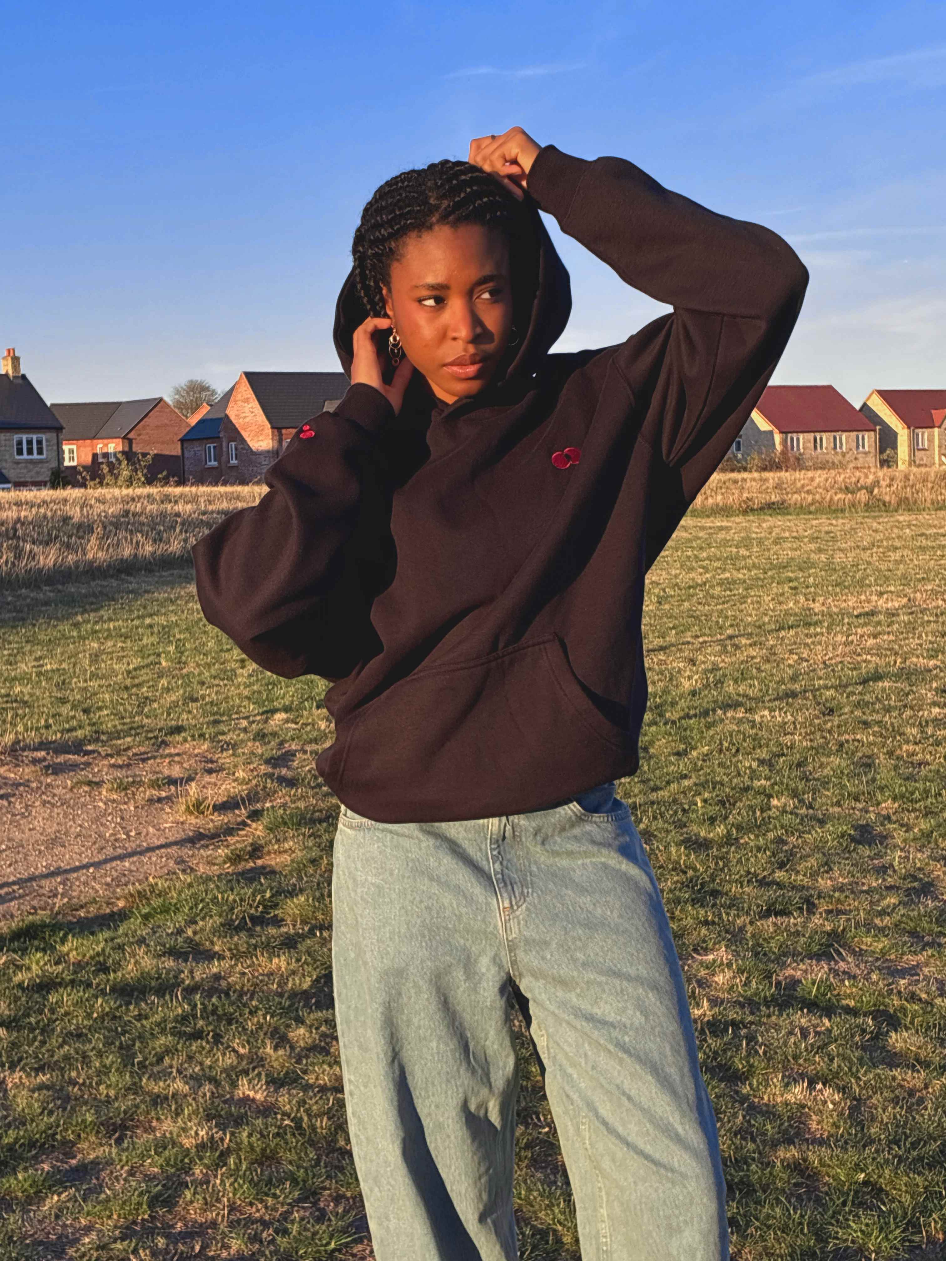 Model pulling up the hood of the black Red Dot Hoodie in warm evening light, the red double-dot on the chest and sleeve, with light-wash wide-leg jeans