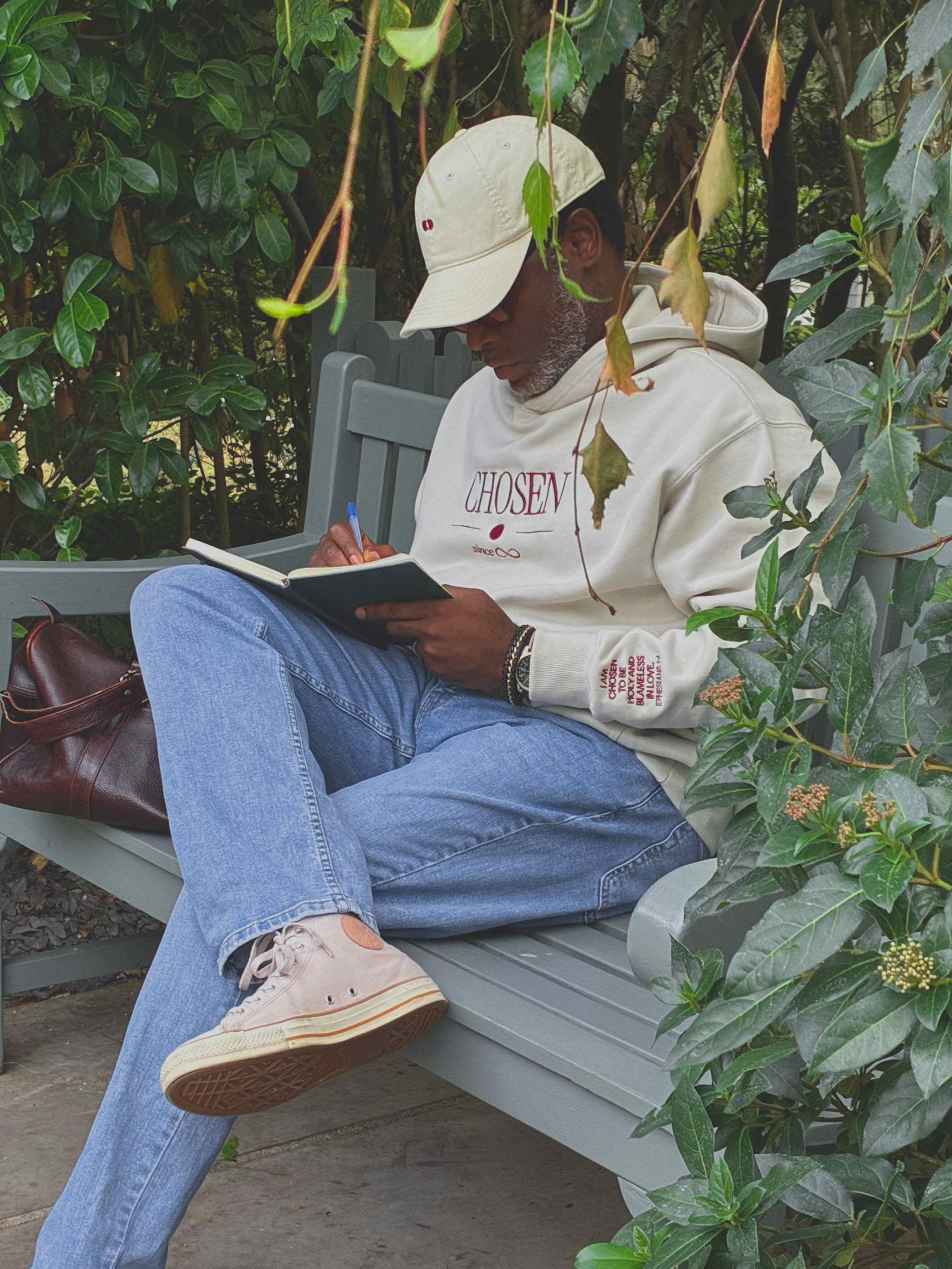 Model writing in a notebook on a garden bench, the Ephesians 1:4 scripture embroidered on the cuff of the cream Chosen hoodie