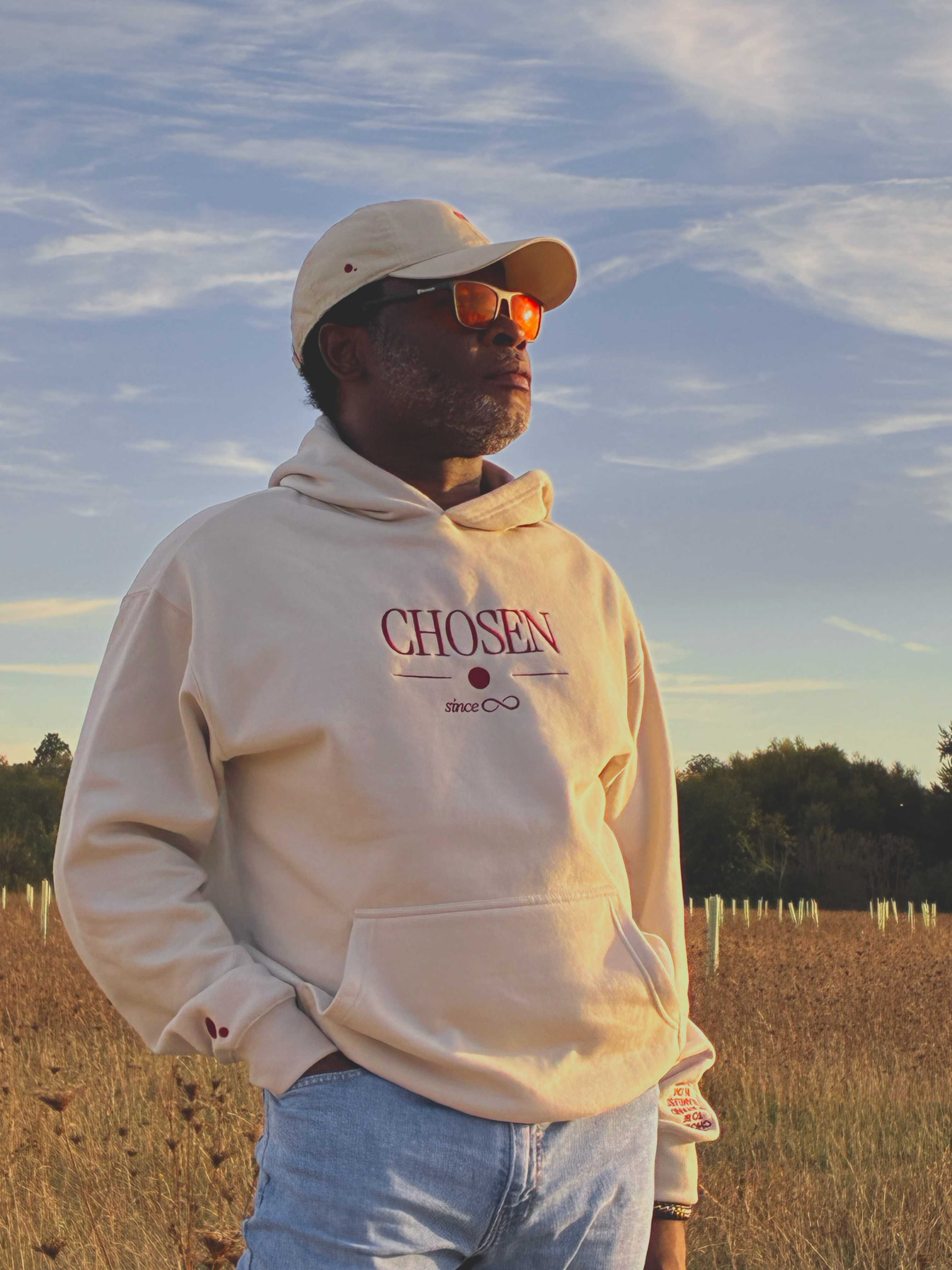 Model in an open field at golden hour, gazing into the distance in the cream Chosen hoodie, Red Dot Cap and orange-tinted sunglasses
