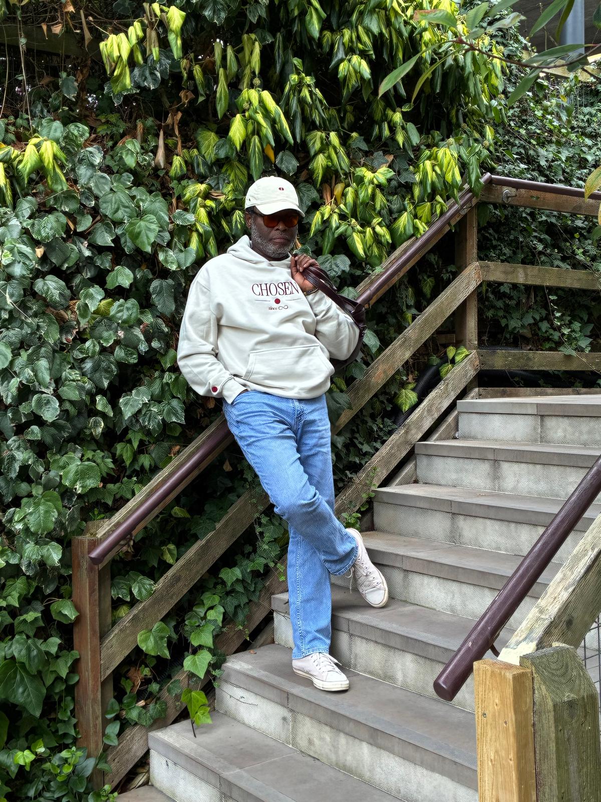 Model leaning on a wooden stair rail against an ivy wall, wearing the cream Chosen hoodie, Red Dot Cap and light-wash jeans, a leather bag over his shoulder