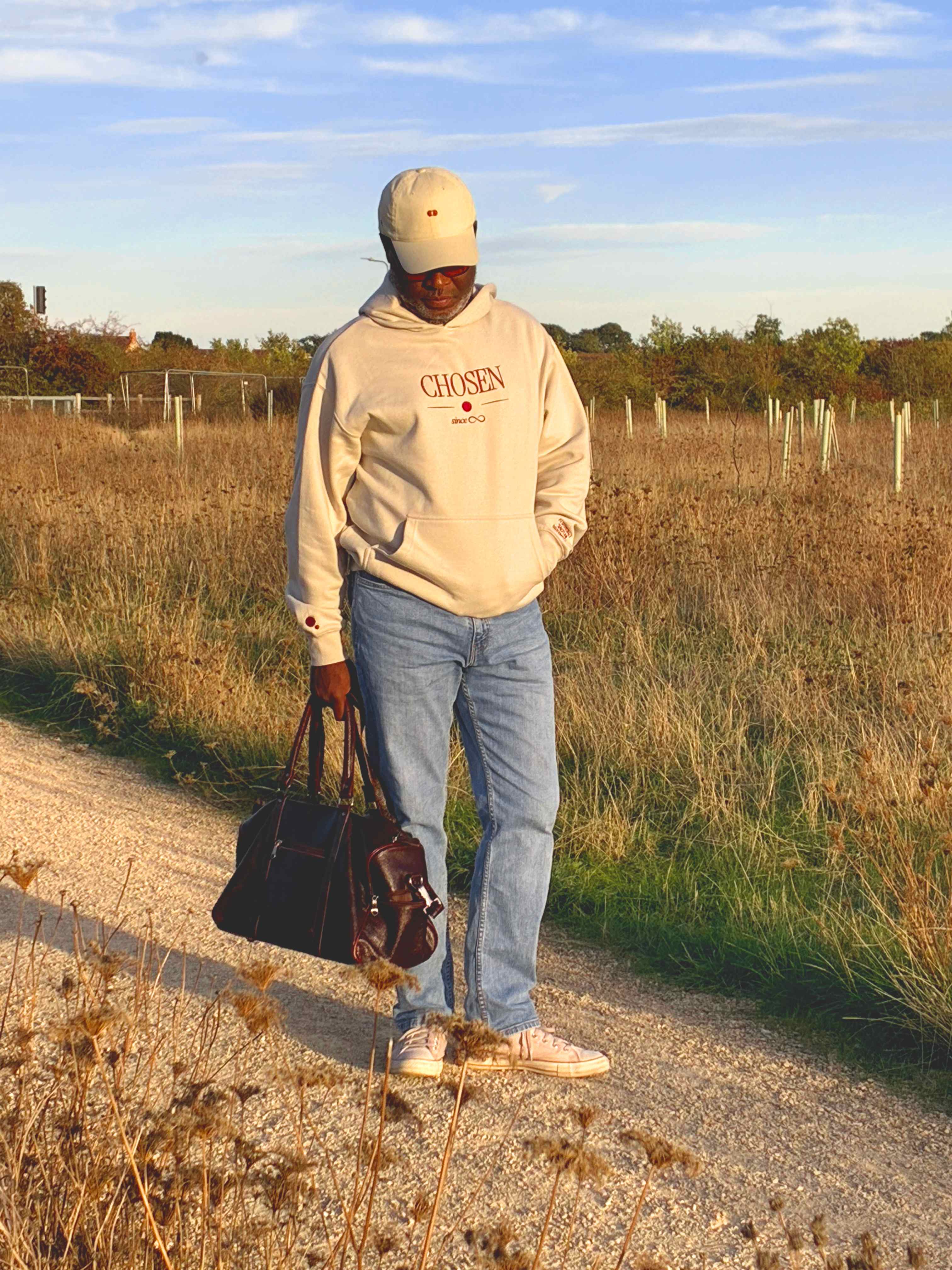 Model on a gravel path through dry grass, looking down, carrying a burgundy leather holdall, in light-wash jeans and pink canvas high-tops