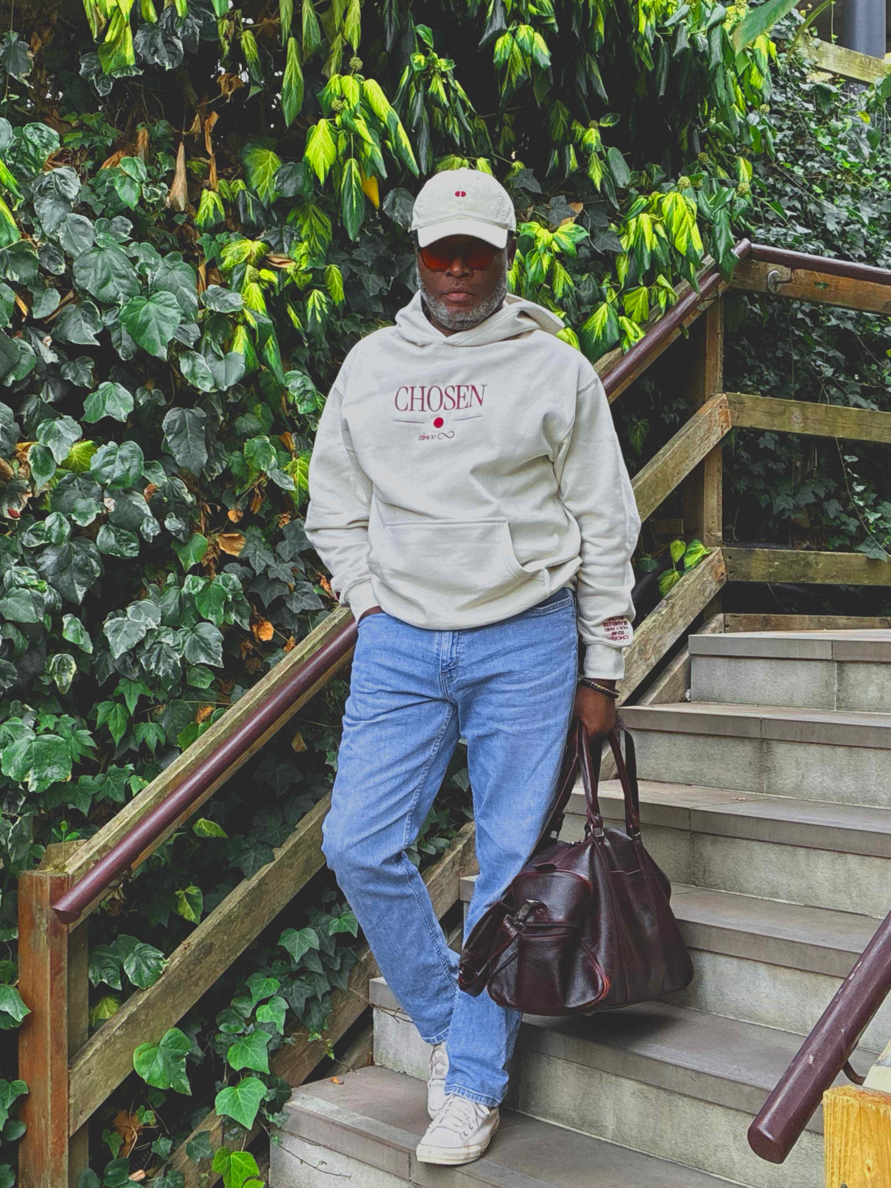Model standing on stone steps against an ivy wall, wearing the cream Chosen hoodie, Red Dot Cap, tinted sunglasses and light-wash jeans, a burgundy leather holdall in hand