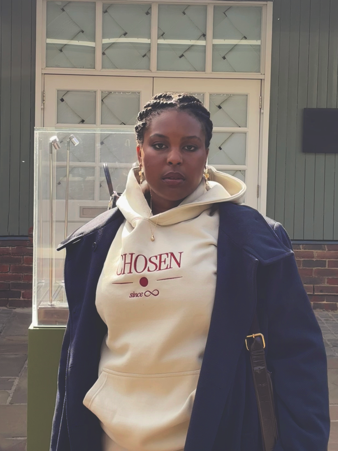 Model facing the camera in the cream Chosen Since Infinity Hoodie and navy coat, the burgundy CHOSEN since infinity embroidery on the chest
