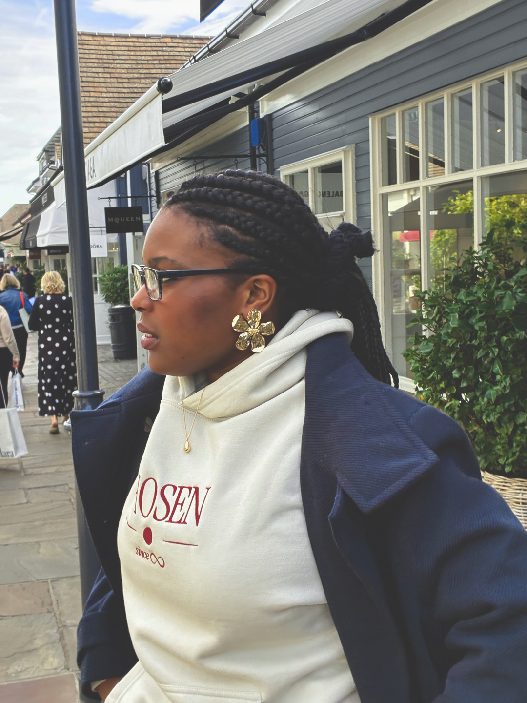 Side profile of the model in glasses and a gold flower earring, wearing the cream Chosen hoodie under a navy coat