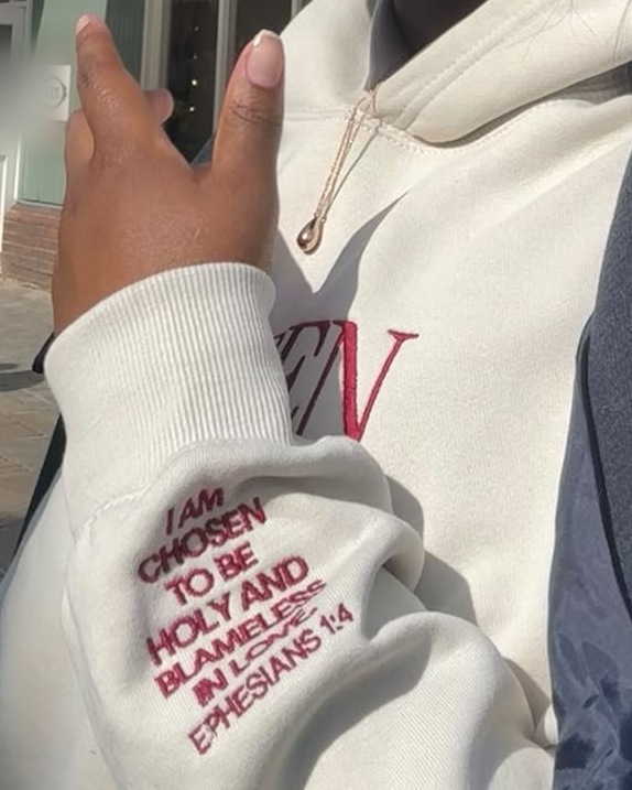 Close-up of the cream Chosen Since Infinity Hoodie sleeve, embroidered in burgundy with "I am chosen to be holy and blameless in love, Ephesians 1:4"