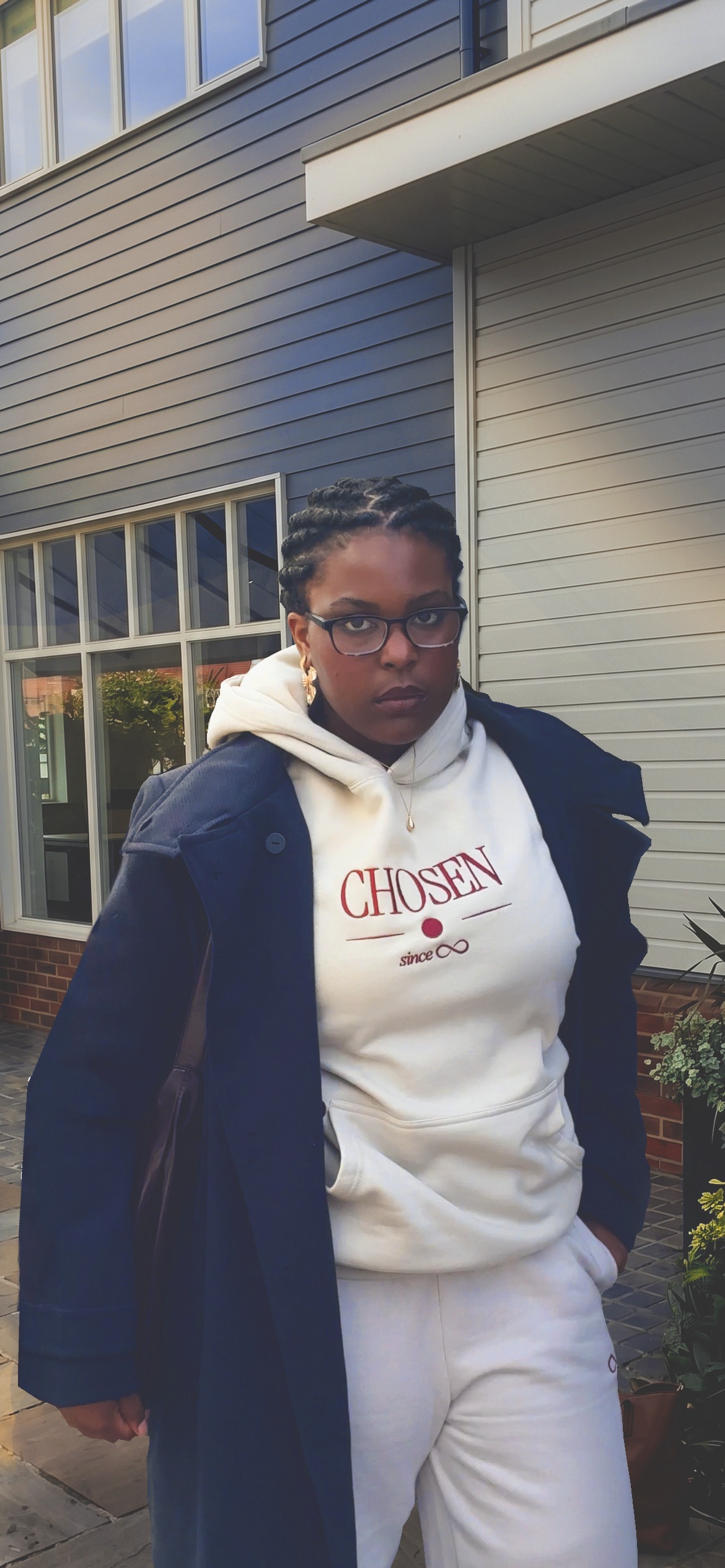 Model in glasses wearing the cream Chosen Since Infinity Hoodie and Sweatpants under an open navy coat, hands in pockets