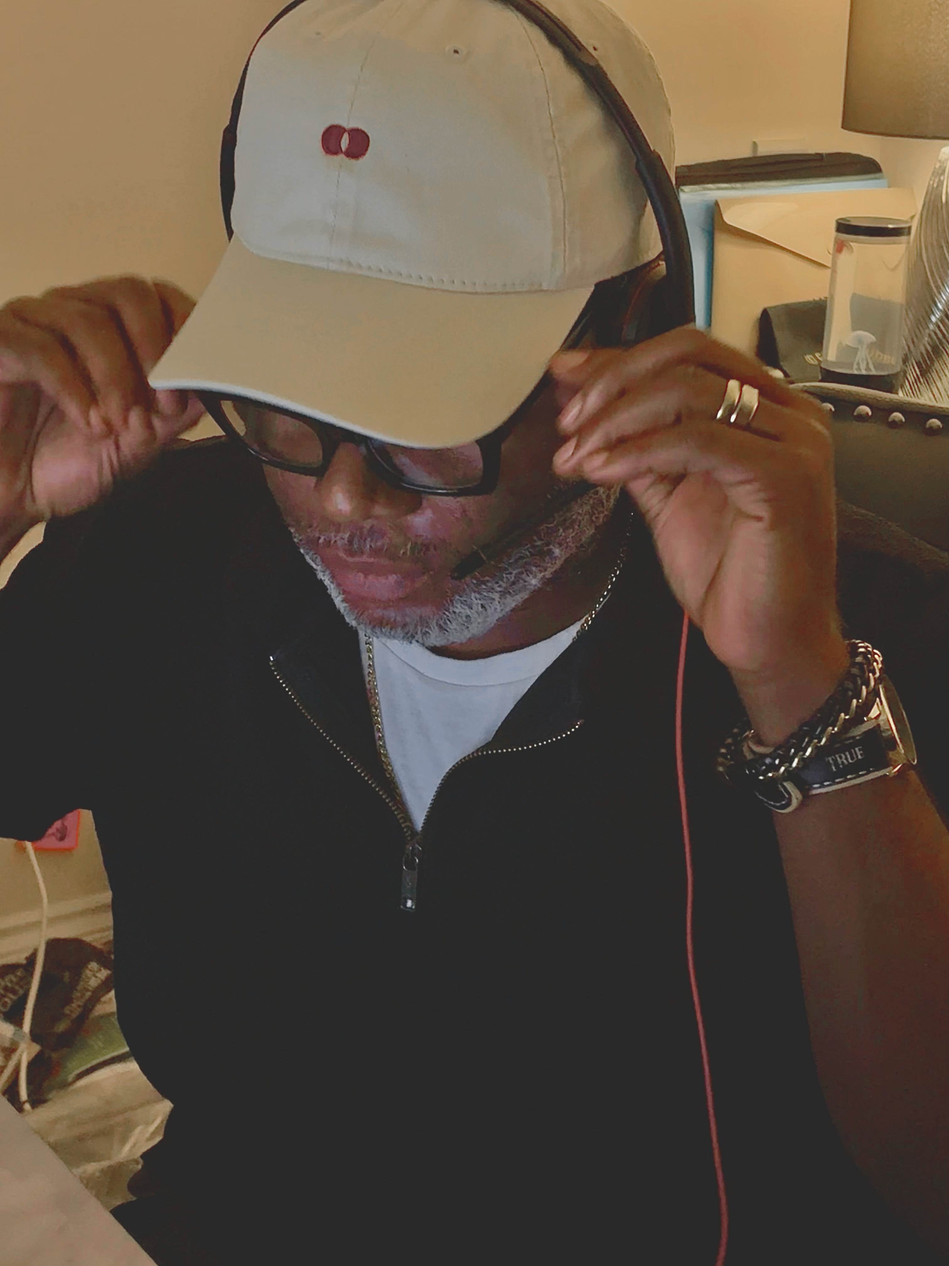 Close-up of the model adjusting the cream Red Dot Cap with both hands, the burgundy double-dot embroidery on the front, a gold ring and a watch strap stamped TRUE