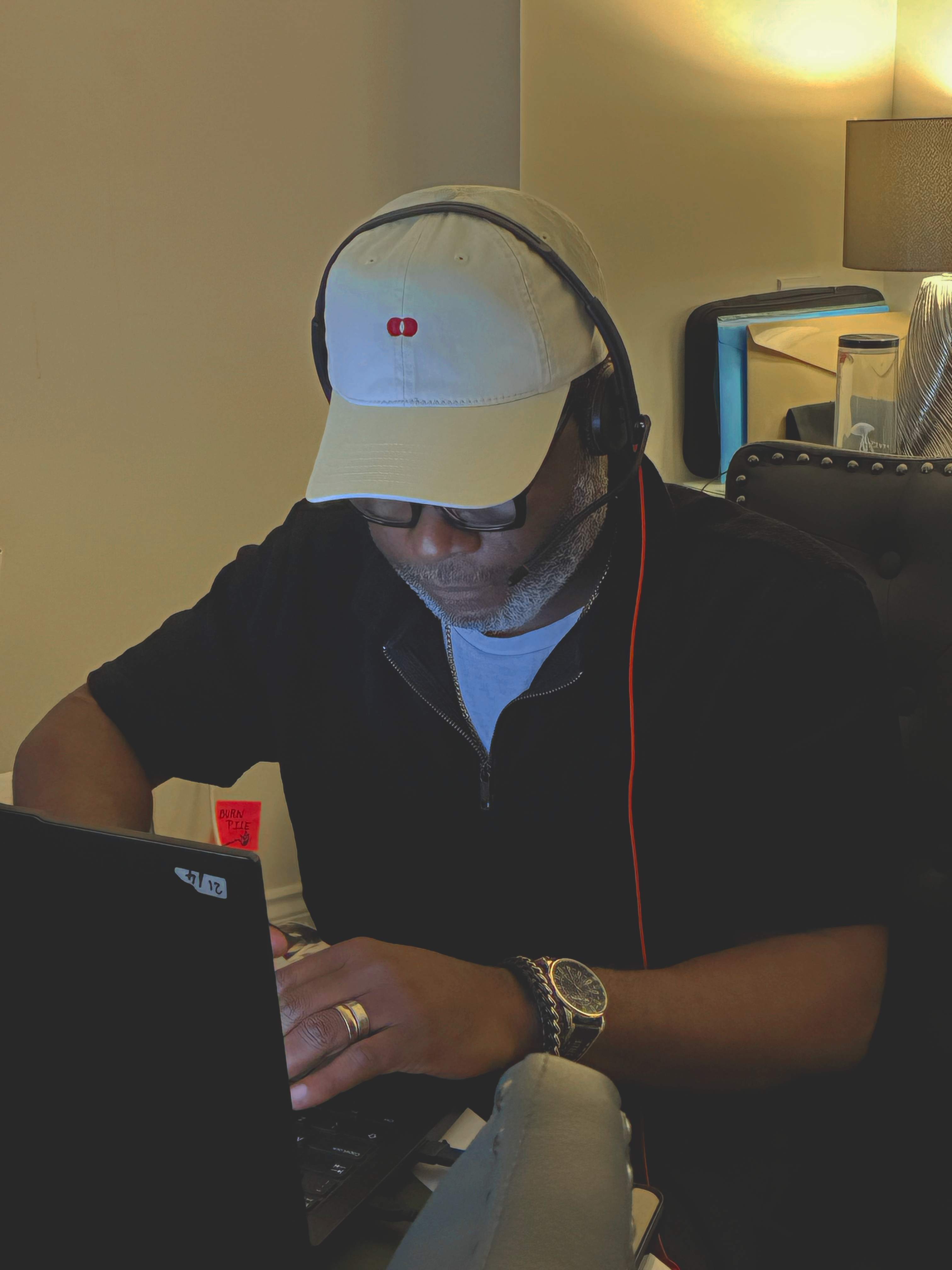 Model in glasses and a headset typing at a laptop, wearing the cream Red Dot Cap and a black quarter-zip