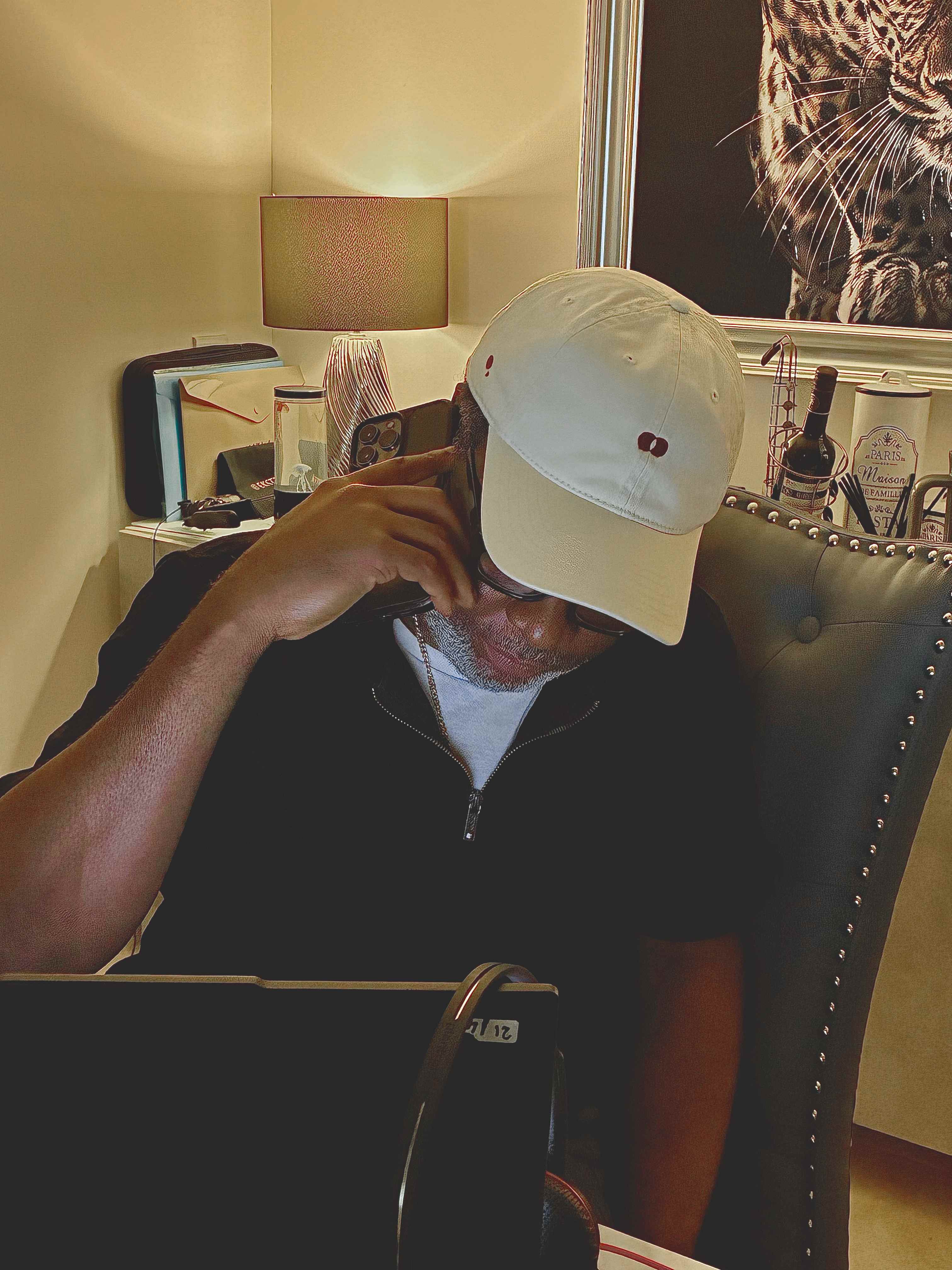 Model on a phone call in a studded leather chair, wearing the cream Red Dot Cap with a black quarter-zip over a white tee, a lamp glowing behind